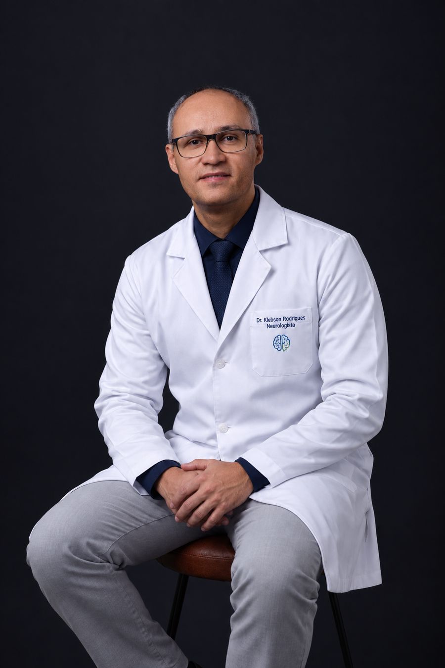 Dr. Klebson Rodrigues sentado, de jaleco e gravata